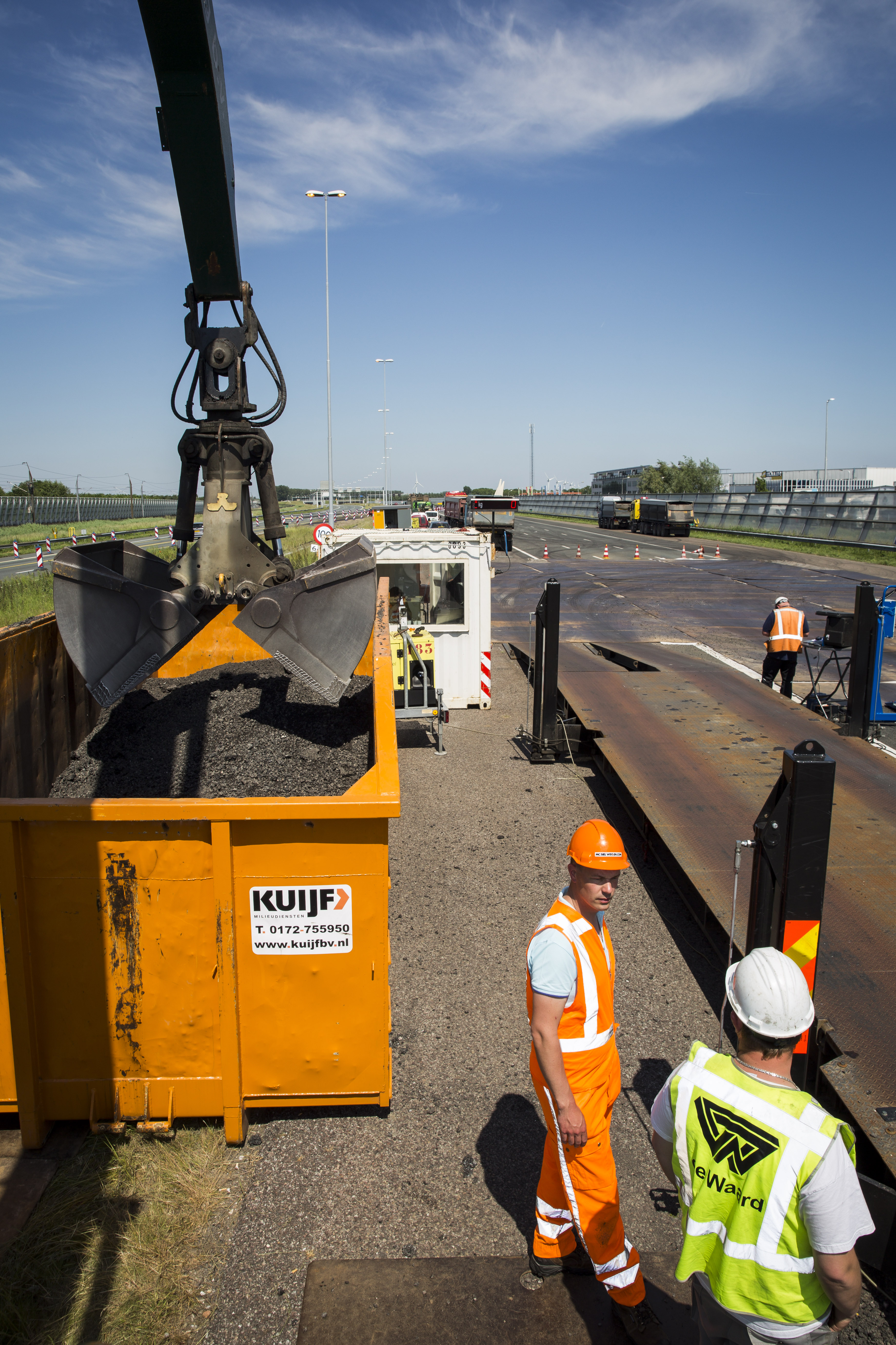 Direct naast de weegbrug staat een container met gefreesd asfalt en een overslagkraan. De kraan haalt materiaal uit de vrachtwagen of laadt bij. Aan de voorzijde van de weegbrug staat een kleine container met daarin de computer.
